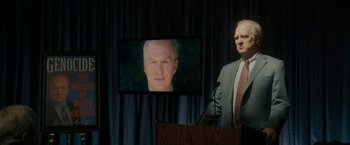 Movie still from “Imperium” (2016), directed by Daniel Ragussis – A man standing at a podium in front of a television; Medium shot, Over the shoulder angle