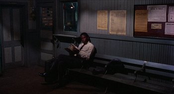 Movie still from “In the Heat of the Night” (1967), directed by Norman Jewison – A man sitting on a bench reading a book; Wide shot, Over the shoulder angle
