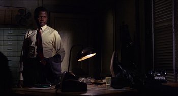 Movie still from “In the Heat of the Night” (1967), directed by Norman Jewison – A man standing in a dimly - lit room next to a desk; Medium shot, Low angle