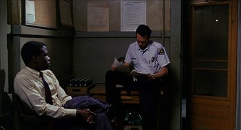 Movie still from “In the Heat of the Night” (1967), directed by Norman Jewison – A police officer sitting on a bench reading a newspaper; Medium shot, High angle