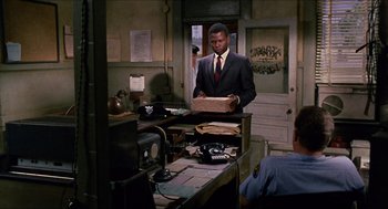 Movie still from “In the Heat of the Night” (1967), directed by Norman Jewison – A man in a suit and tie holding a box in front of a desk; Medium shot, Over the shoulder angle