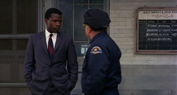 Movie still from “In the Heat of the Night” (1967), directed by Norman Jewison – A man in a suit and tie standing next to a man in a police uniform; Medium shot, Over the shoulder angle