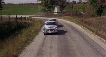 Movie still from “In the Heat of the Night” (1967), directed by Norman Jewison – A couple of cars driving down a road near a field; Extreme Wide shot, High angle