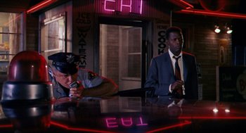 Movie still from “In the Heat of the Night” (1967), directed by Norman Jewison – Two men sitting at a table in front of neon lights; Wide shot, Over the shoulder angle