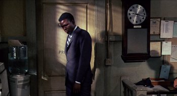 Movie still from “In the Heat of the Night” (1967), directed by Norman Jewison – A man in a suit and tie standing in front of a clock; Medium shot, High angle