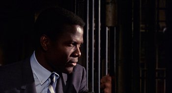 Movie still from “In the Heat of the Night” (1967), directed by Norman Jewison – A man in a suit and tie sitting in front of a metal fence; Close Up shot, Low angle