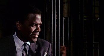 Movie still from “In the Heat of the Night” (1967), directed by Norman Jewison – A man wearing a suit and tie sitting in front of a metal fence; Close Up shot, Over the shoulder angle