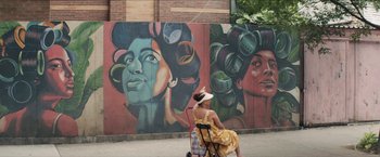 Movie still from “In the Heights” (2021), directed by Jon M. Chu – A woman sitting on a chair in front of a mural; Wide shot, Low angle