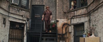 Movie still from “In the Heights” (2021), directed by Jon M. Chu – A man in a brown suit and hat standing on a set of stairs; Wide shot, Low angle