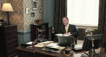 Movie still from “In the Loop” (2009), directed by Armando Iannucci – An older man sitting at a desk in front of an open laptop computer; Wide shot, Over the shoulder angle