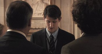 Movie still from “In the Loop” (2009), directed by Armando Iannucci – A man in a suit and tie standing next to two other men; Close Up shot, Over the shoulder angle