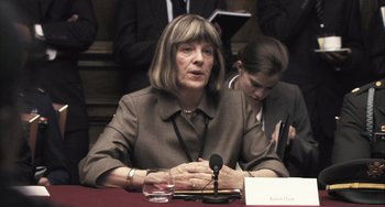 Movie still from “In the Loop” (2009), directed by Armando Iannucci – An older woman sitting in front of a microphone at a table; Close Up shot, Over the shoulder angle