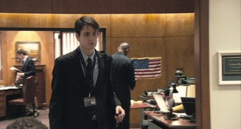 Movie still from “In the Loop” (2009), directed by Armando Iannucci – A man in a suit and tie standing in front of an american flag; Medium shot, Over the shoulder angle