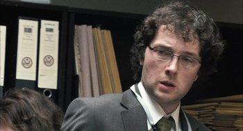 Movie still from “In the Loop” (2009), directed by Armando Iannucci – A man in a suit and tie in front of a book case; Close Up shot, Over the shoulder angle