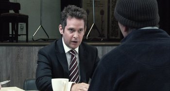 Movie still from “In the Loop” (2009), directed by Armando Iannucci – A man wearing a suit and tie holding a cup of coffee talking to another man wearing a suit and tie; Medium shot, Over the shoulder angle
