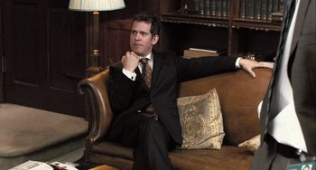 Movie still from “In the Loop” (2009), directed by Armando Iannucci – A man in a suit and tie sitting on a couch; Medium shot, Over the shoulder angle