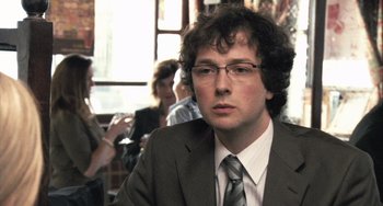 Movie still from “In the Loop” (2009), directed by Armando Iannucci – A man in a suit and tie sitting in front of other people; Close Up shot, Over the shoulder angle