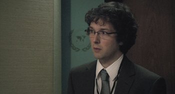 Movie still from “In the Loop” (2009), directed by Armando Iannucci – A man in a suit and tie standing in front of a wall; Close Up shot, Over the shoulder angle