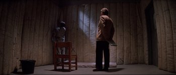 Movie still from “In the Mouth of Madness” (1994), directed by John Carpenter – Two people standing in front of a curtain and a chair; Wide shot, Over the shoulder angle