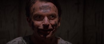 Movie still from “In the Mouth of Madness” (1994), directed by John Carpenter – A man with tattoos on his face smiles for the camera; Close Up shot, Over the shoulder angle