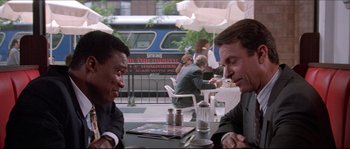 Movie still from “In the Mouth of Madness” (1994), directed by John Carpenter – Two men sitting at a table in front of an umbrella; Medium shot, Over the shoulder angle