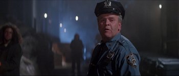 Movie still from “In the Mouth of Madness” (1994), directed by John Carpenter – A man in a police uniform standing next to a building; Medium shot, Low angle