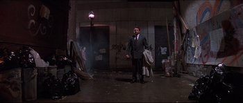 Movie still from “In the Mouth of Madness” (1994), directed by John Carpenter – A man wearing a suit and tie standing in a room; Wide shot, Low angle