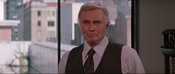 Movie still from “In the Mouth of Madness” (1994), directed by John Carpenter – An older man wearing a vest and a tie; Medium shot, Over the shoulder angle