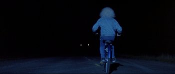 Movie still from “In the Mouth of Madness” (1994), directed by John Carpenter – A person riding a bike at night on the street; Wide shot, Over the shoulder angle