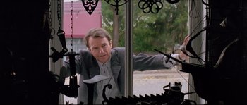 Movie still from “In the Mouth of Madness” (1994), directed by John Carpenter – A man in a suit and tie leaning against a window; Medium shot, Low angle