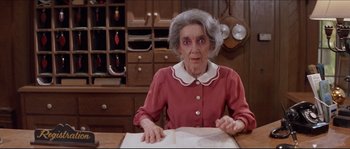 Movie still from “In the Mouth of Madness” (1994), directed by John Carpenter – An older woman sitting at a table with a book; Medium shot, High angle