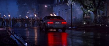 Movie still from “In the Mouth of Madness” (1994), directed by John Carpenter – A car driving down a street at night with people walking around; Wide shot, High angle