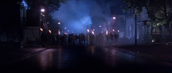 Movie still from “In the Mouth of Madness” (1994), directed by John Carpenter – A group of people standing on a street with torches; Extreme Wide shot, High angle
