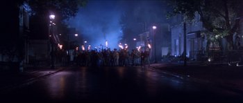 Movie still from “In the Mouth of Madness” (1994), directed by John Carpenter – A group of people standing on the side of the road holding torches; Extreme Wide shot, High angle