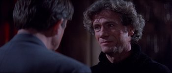 Movie still from “In the Mouth of Madness” (1994), directed by John Carpenter – A man in a black shirt is talking to another man; Close Up shot, Over the shoulder angle
