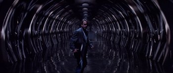 Movie still from “In the Mouth of Madness” (1994), directed by John Carpenter – A man in a suit walking through a tunnel; Wide shot, Low angle