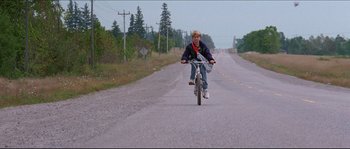 Movie still from “In the Mouth of Madness” (1994), directed by John Carpenter – A man riding a bike down the middle of the road; Wide shot, Low angle