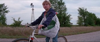 Movie still from “In the Mouth of Madness” (1994), directed by John Carpenter – A young boy riding a bike with a globe bag on the back of it; Medium shot, Low angle