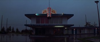 Movie still from “In the Mouth of Madness” (1994), directed by John Carpenter – A motel sign lit up at night in the rain; Extreme Wide shot, Low angle