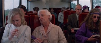 Movie still from “In the Mouth of Madness” (1994), directed by John Carpenter – An older woman sitting on a train holding a phone; Medium shot, Over the shoulder angle