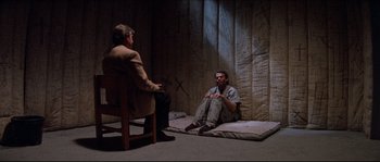 Movie still from “In the Mouth of Madness” (1994), directed by John Carpenter – Two men sitting in a dimly - lit room talking; Wide shot, High angle