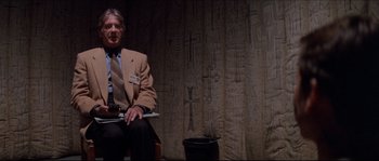 Movie still from “In the Mouth of Madness” (1994), directed by John Carpenter – An older man in a suit and tie sitting on a chair; Medium shot, Low angle