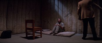 Movie still from “In the Mouth of Madness” (1994), directed by John Carpenter – A man sitting on the floor in a room; Wide shot, High angle