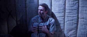 Movie still from “In the Mouth of Madness” (1994), directed by John Carpenter – A man in a hospital gown holding a pair of gloves; Medium shot, Overhead angle