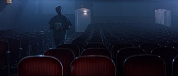 Movie still from “In the Mouth of Madness” (1994), directed by John Carpenter – A man standing in front of rows of empty chairs; Wide shot, High angle