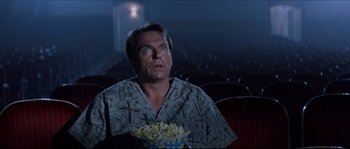 Movie still from “In the Mouth of Madness” (1994), directed by John Carpenter – A man sitting in front of a bowl of popcorn in a movie theater; Medium shot, Low angle