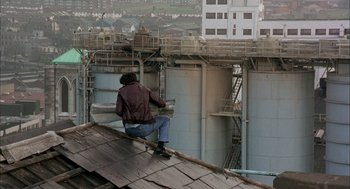 Movie still from “In the Name of the Father” (1993), directed by Jim Sheridan – A man sitting on the roof of a building on a skateboard; Wide shot, High angle