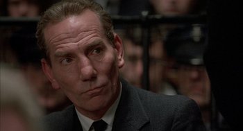 Movie still from “In the Name of the Father” (1993), directed by Jim Sheridan – A man in a suit and tie sitting in front of a group of people; Close Up shot, Low angle