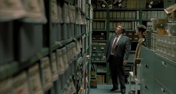 Movie still from “Incendies” (2010), directed by Denis Villeneuve – A man in a suit and tie looking up at a wall of files; Medium shot, Over the shoulder angle