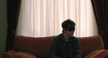 Movie still from “Incendies” (2010), directed by Denis Villeneuve – A man sitting on top of a couch wearing a blindfold; Medium shot, Over the shoulder angle